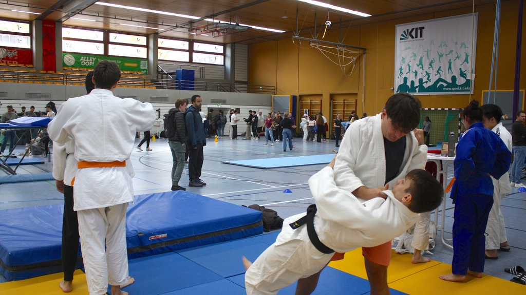 Judo-Training in einer Sporthalle mit mehreren Paaren und Zuschauern im Hintergrund.