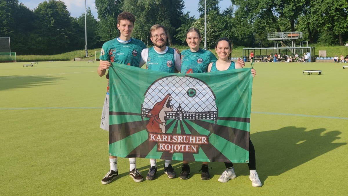 Vier Personen halten eine Flagge der "Karlsruher Koyoten" auf einem Spielfeld.